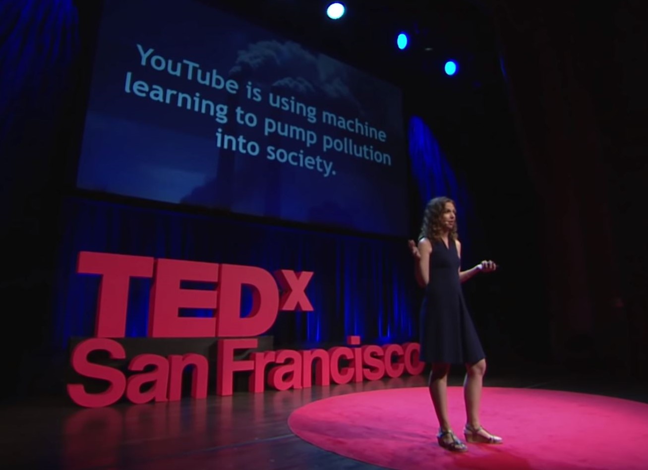 Women in a navy dress standing onstage with a red sign saying TEDx San Francisco behind her. Slide is visible with words "YouTube is using machine learning to pump pollution into society"